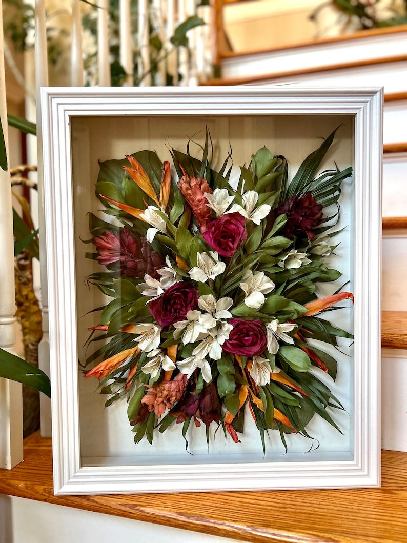 May include: A framed pressed flower arrangement with a variety of dried flowers and leaves, including red roses, white flowers, orange and green leaves, and brown ginger flowers.