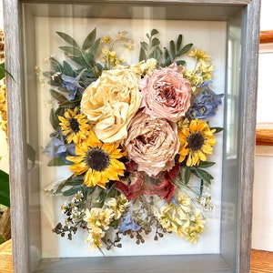 May include: A framed pressed flower arrangement with sunflowers, roses, and other dried flowers in shades of yellow, pink, and blue.
