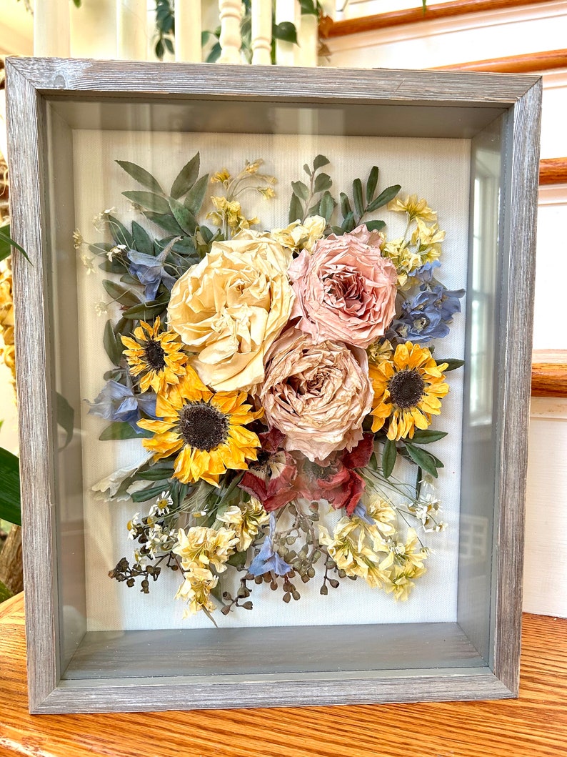 May include: A framed pressed flower arrangement with sunflowers, roses, and other dried flowers in shades of yellow, pink, and blue.