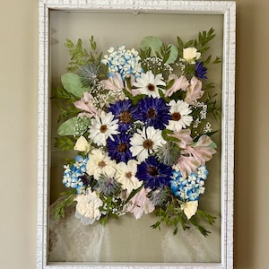 May include: A rectangular white distressed wooden frame containing a pressed flower arrangement. The display features deep blue cornflowers, white daisies, light blue hydrangea florets, green eucalyptus leaves, and small thistle accents.