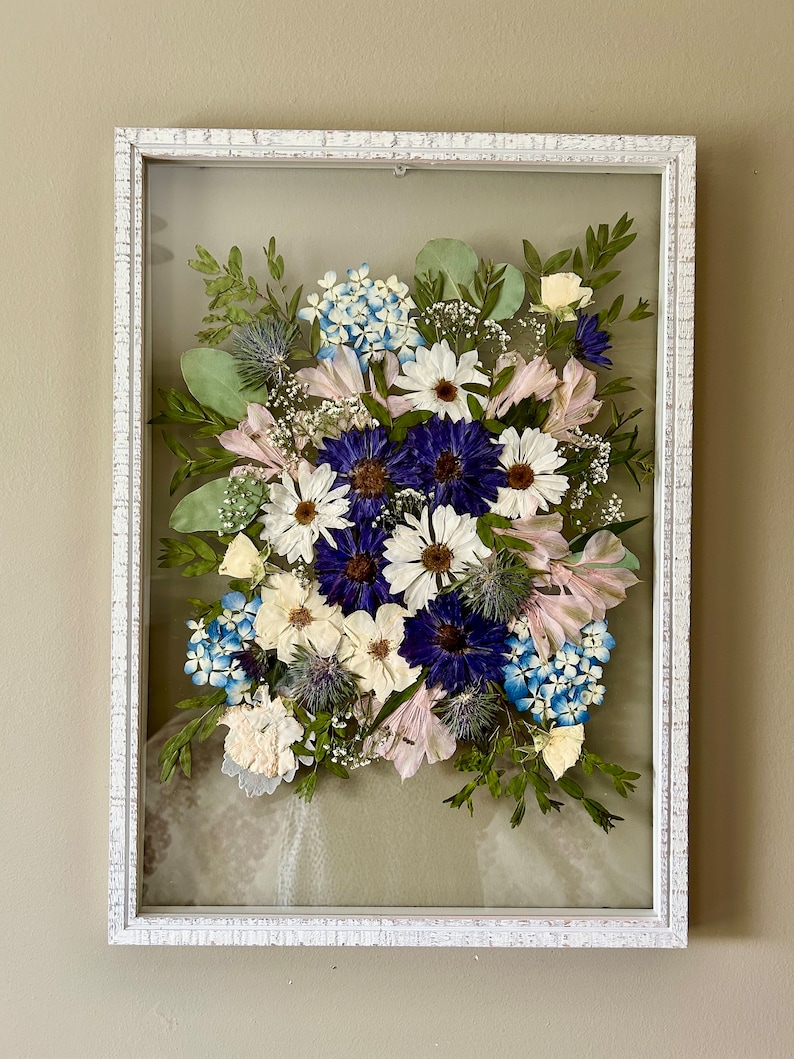 May include: A rectangular white distressed wooden frame containing a pressed flower arrangement. The display features deep blue cornflowers, white daisies, light blue hydrangea florets, green eucalyptus leaves, and small thistle accents.