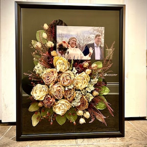 May include: A framed pressed flower arrangement with a black frame and a photo of a couple in the background. The flowers are mostly beige and brown with some red accents.
