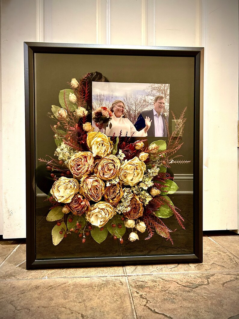 May include: A framed pressed flower arrangement with a black frame and a photo of a couple in the background. The flowers are mostly beige and brown with some red accents.