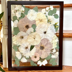 May include: A rectangular dark brown frame containing a pressed floral arrangement. The display features various dried flowers in shades of cream, pale pink, and muted mauve, interspersed with small green eucalyptus leaves against a clear background.