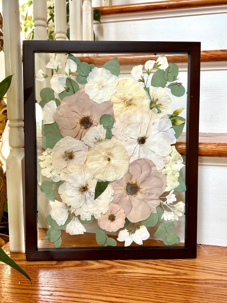 May include: A rectangular dark brown frame containing a pressed floral arrangement. The display features various dried flowers in shades of cream, pale pink, and muted mauve, interspersed with small green eucalyptus leaves against a clear background.