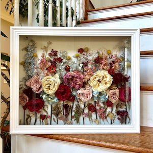 May include: A white frame with a glass front displays a pressed flower arrangement. The flowers are a mix of pink, white, red, and yellow, with a variety of greenery. The arrangement is set against a light beige background.