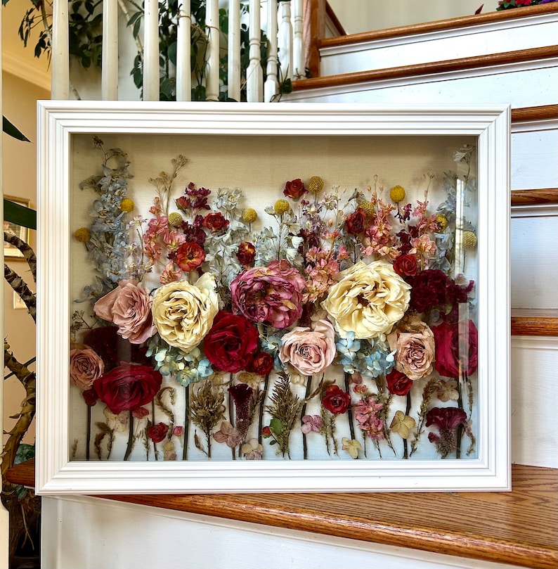 May include: A white frame with a glass front displays a pressed flower arrangement. The flowers are a mix of pink, white, red, and yellow, with a variety of greenery. The arrangement is set against a light beige background.