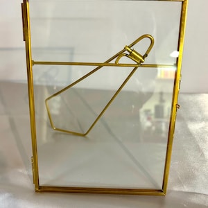 May include: A rectangular, gold-coloured metal picture frame with a clear glass pane. The frame has a hinged back and a gold-coloured metal stand. The frame is designed to hold a photograph or artwork.