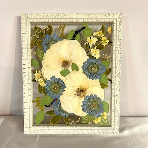 May include: A rectangular white distressed wooden frame containing pressed flowers. The arrangement features two large cream-colored blossoms, several smaller blue flowers, green leaves, and delicate yellow sprigs preserved between two clear glass panes.