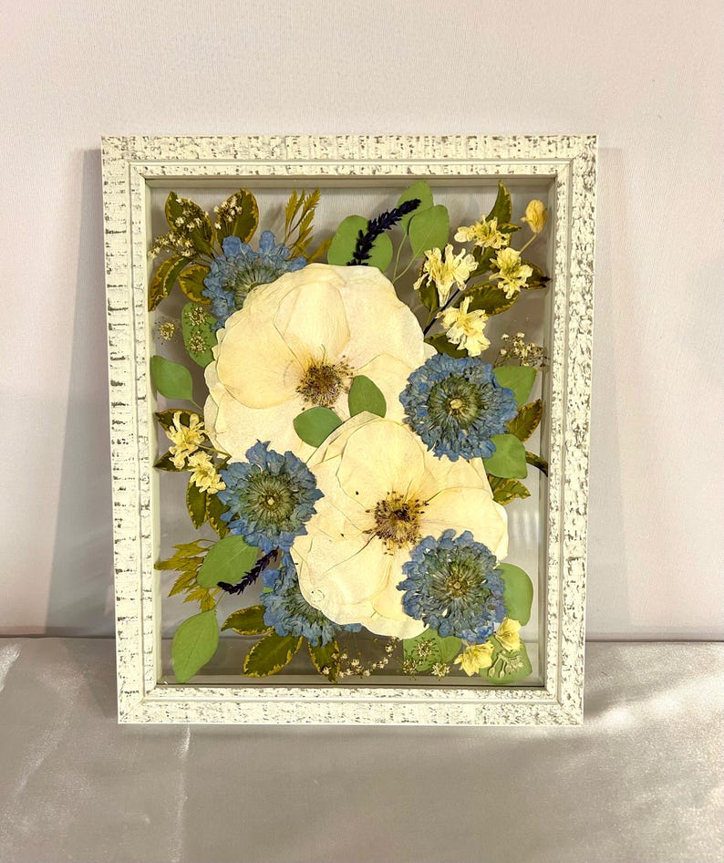 May include: A rectangular white distressed wooden frame containing pressed flowers. The arrangement features two large cream-colored blossoms, several smaller blue flowers, green leaves, and delicate yellow sprigs preserved between two clear glass panes.