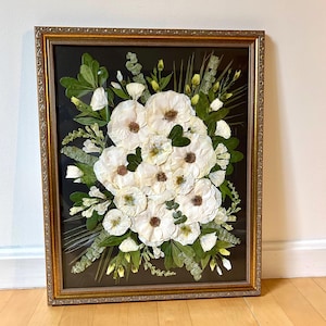 May include: A rectangular wall art piece featuring pressed white flowers and green foliage arranged on a black background. The arrangement is enclosed in a decorative gold-toned frame with an ornate, textured border and a clear glass front panel.