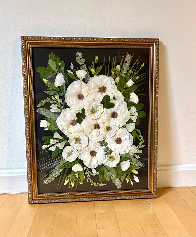May include: A rectangular wall art piece featuring pressed white flowers and green foliage arranged on a black background. The arrangement is enclosed in a decorative gold-toned frame with an ornate, textured border and a clear glass front panel.