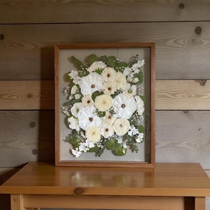May include: A rectangular wooden frame containing a pressed floral arrangement featuring white and cream-colored flowers, green eucalyptus leaves, and small white filler flowers set against a neutral background within the glass display.