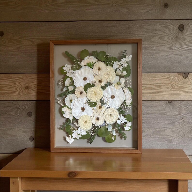 May include: A rectangular wooden frame containing a pressed floral arrangement featuring white and cream-colored flowers, green eucalyptus leaves, and small white filler flowers set against a neutral background within the glass display.