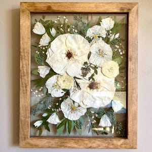 May include: A rectangular wooden frame containing a pressed floral arrangement featuring large white flowers with dark centers, smaller white blossoms, and various green leaves and sprigs of baby's breath preserved behind a clear glass pane.
