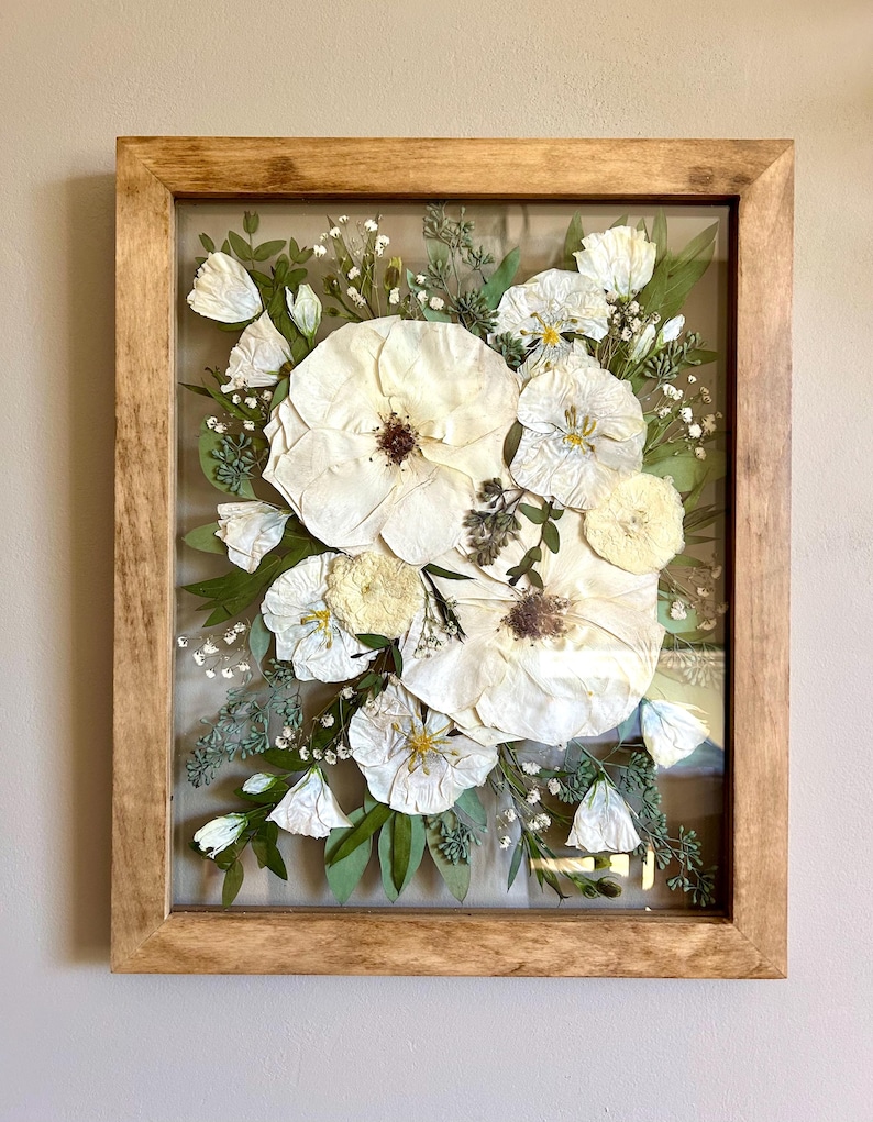 May include: A rectangular wooden frame containing a pressed floral arrangement featuring large white flowers with dark centers, smaller white blossoms, and various green leaves and sprigs of baby's breath preserved behind a clear glass pane.