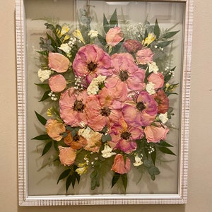May include: A rectangular wall frame with a distressed white finish containing a pressed flower arrangement. The display features pink and peach-toned flowers, small white blossoms, and green foliage set against a neutral background.