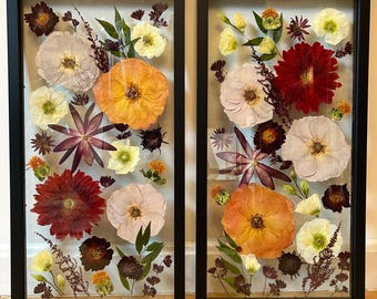 May include: Two black framed rectangular shadow boxes with pressed flowers and greenery. The flowers are in shades of pink, red, orange, yellow, and white. The greenery is in shades of green and brown.