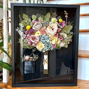 May include: A preserved wedding bouquet in a black frame with a white ribbon and a gold letter "K" on the ribbon. The bouquet includes roses, hydrangeas, and other flowers in shades of pink, yellow, and blue.