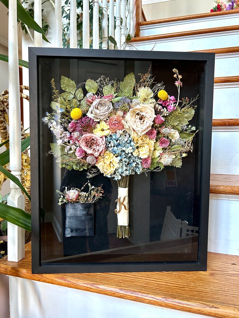 May include: A preserved wedding bouquet in a black frame with a white ribbon and a gold letter "K" on the ribbon. The bouquet includes roses, hydrangeas, and other flowers in shades of pink, yellow, and blue.