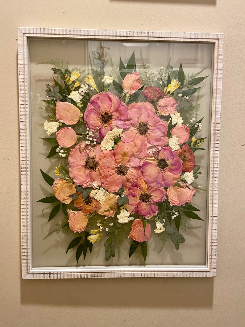 May include: A rectangular wall frame with a distressed white finish containing a pressed flower arrangement. The display features pink and peach-toned flowers, small white blossoms, and green foliage set against a neutral background.