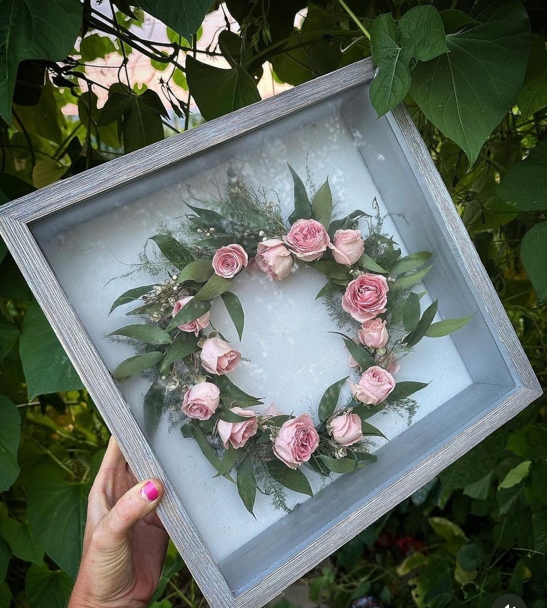 A gray wooden shadow box with a glass front containing a preserved bridal crown . Boutonnière, corsage etc. can also be preserved like this.