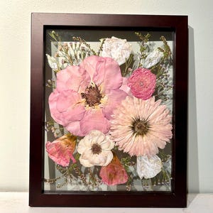 May include: A rectangular dark brown wooden frame containing a pressed flower arrangement. The display features a large central pink flower, a light pink daisy-like bloom, smaller white and pink blossoms, and delicate green foliage behind glass.