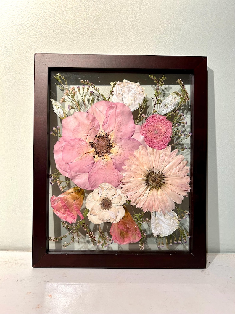 May include: A rectangular dark brown wooden frame containing a pressed flower arrangement. The display features a large central pink flower, a light pink daisy-like bloom, smaller white and pink blossoms, and delicate green foliage behind glass.