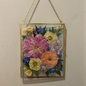 May include: A rectangular glass frame with a thin gold metal border, suspended by a gold chain. Inside the frame are pressed flowers in shades of pink, peach, yellow, blue, and purple, arranged with green leaves against a light gray background.
