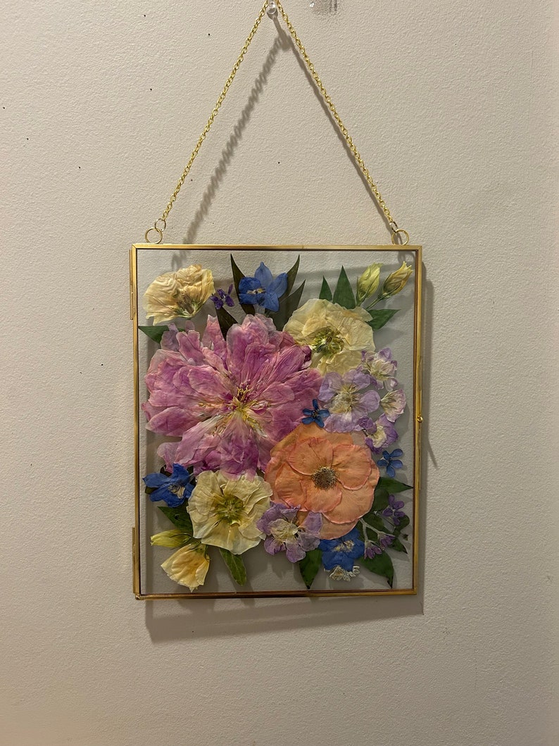 May include: A rectangular glass frame with a thin gold metal border, suspended by a gold chain. Inside the frame are pressed flowers in shades of pink, peach, yellow, blue, and purple, arranged with green leaves against a light gray background.