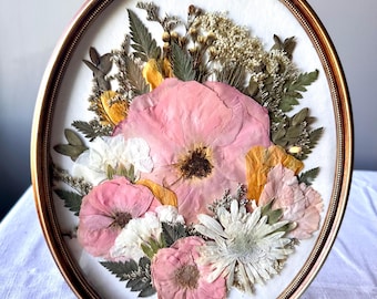 May include: An oval wall frame with a metallic gold finish and a decorative rose-shaped crest at the top. Inside the frame, a collection of pressed dried flowers in shades of pink, white, and yellow are arranged with green fern leaves.