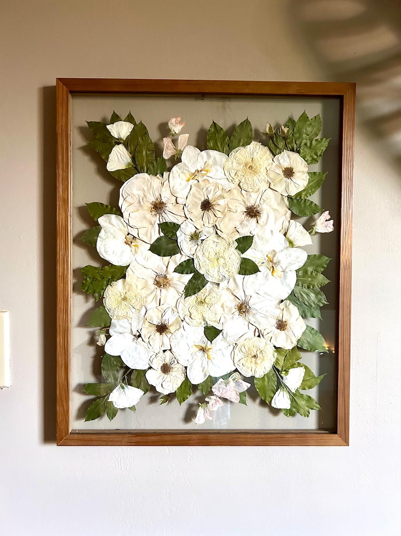May include: A rectangular wooden frame containing a pressed floral arrangement. The display features numerous white flowers with visible brown centers and green leaves, all set against a neutral background behind a clear glass pane.