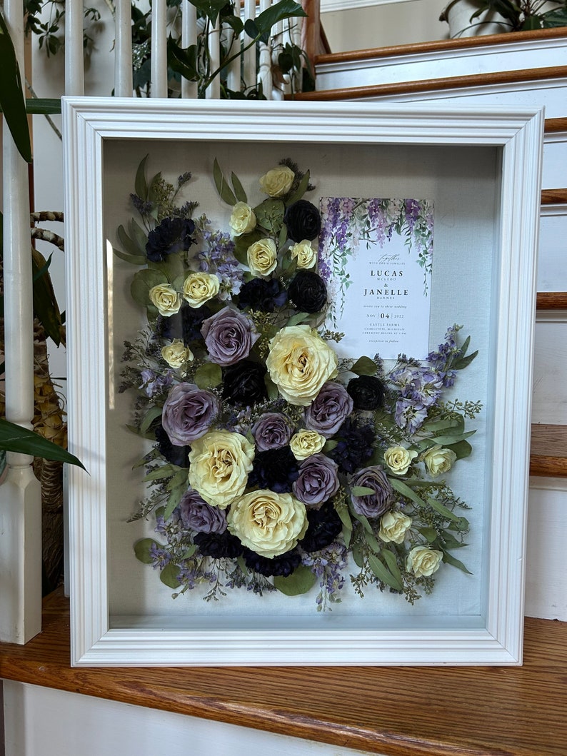 May include: A white framed shadow box with a pressed flower arrangement. The flowers are in shades of purple, yellow, and green. A wedding invitation is visible in the background. The invitation reads "Together with our families, we invite you to celebrate the marriage of Lucas and Janelle."