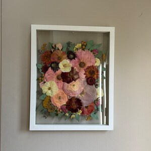 May include: A rectangular white frame containing a pressed floral arrangement. The display features a variety of dried flowers in shades of pink, deep red, yellow, and muted green, including large petals and small buds arranged behind glass.