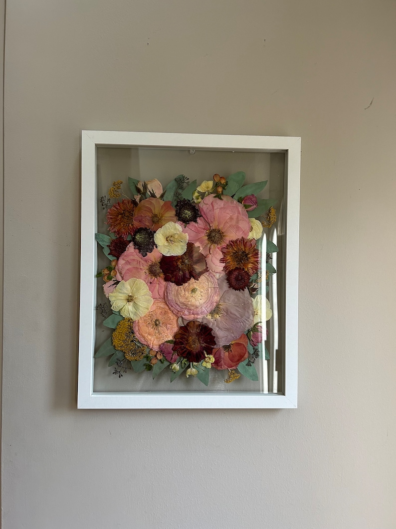 May include: A rectangular white frame containing a pressed floral arrangement. The display features a variety of dried flowers in shades of pink, deep red, yellow, and muted green, including large petals and small buds arranged behind glass.