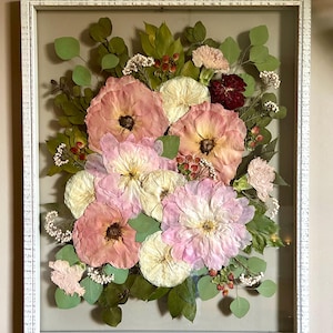 May include: A rectangular white distressed wood frame containing a pressed floral arrangement. The display features various dried flowers in shades of pink, cream, and deep red, accented with green eucalyptus leaves and small white filler flowers.