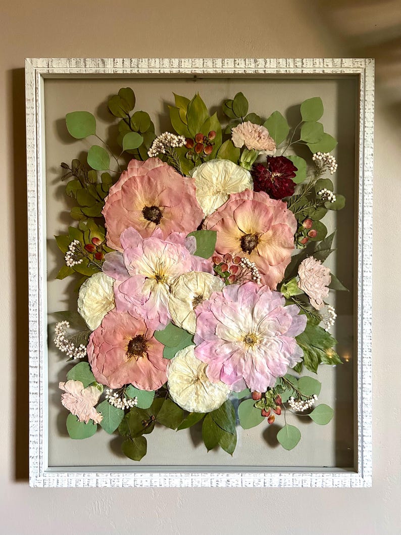 May include: A rectangular white distressed wood frame containing a pressed floral arrangement. The display features various dried flowers in shades of pink, cream, and deep red, accented with green eucalyptus leaves and small white filler flowers.