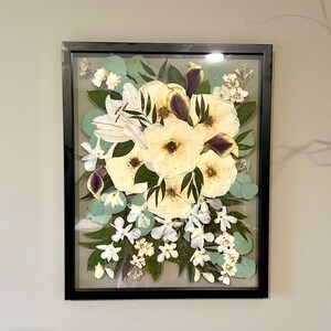 May include: A rectangular black frame containing a pressed floral arrangement. The display features large cream-colored flowers with dark centers, interspersed with green eucalyptus leaves, small white blossoms, and deep purple petal accents.