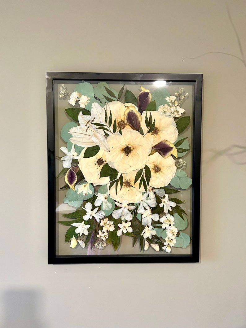 May include: A rectangular black frame containing a pressed floral arrangement. The display features large cream-colored flowers with dark centers, interspersed with green eucalyptus leaves, small white blossoms, and deep purple petal accents.