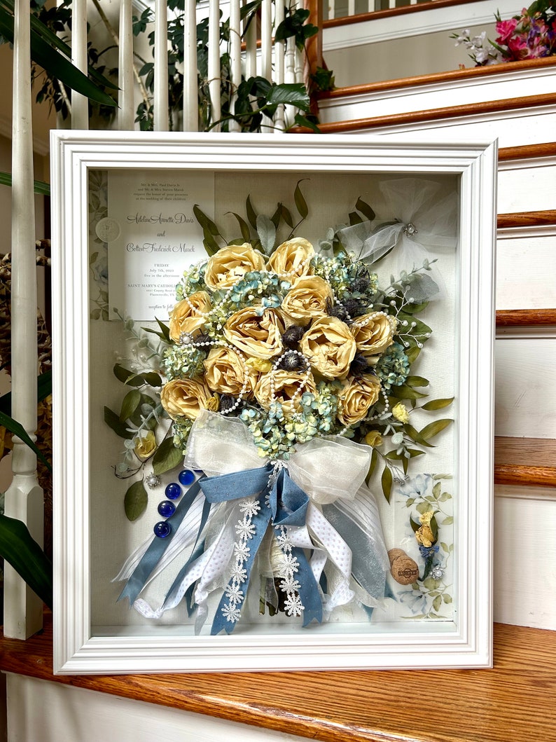 May include: A white framed shadow box with a preserved wedding bouquet .The shadow box also includes a wedding invitation, Boutonniere and original charms and ribbons!