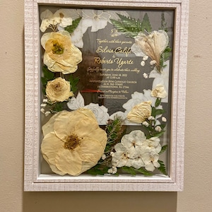 May include: A white textured frame containing a wedding invitation for Silvia Calife and Roberto Ugarte, dated June 18, 2022. The invitation is surrounded by pressed cream-colored flowers, green leaves, and small white baby's breath accents.