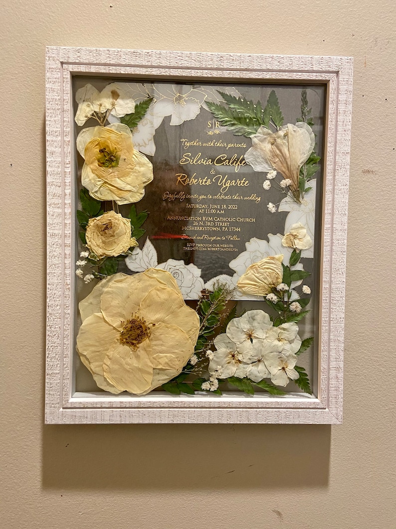 May include: A white textured frame containing a wedding invitation for Silvia Calife and Roberto Ugarte, dated June 18, 2022. The invitation is surrounded by pressed cream-colored flowers, green leaves, and small white baby's breath accents.