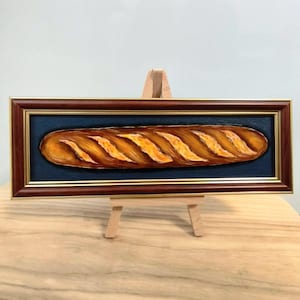 May include: A framed painting of a loaf of bread. The bread is a golden brown colour with diagonal yellow-orange slashes across the top. The painting is set against a dark blue background and framed in a dark brown frame.