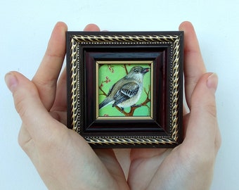 May include: A small framed painting depicting a bird perched on a branch with red berries. The bird is painted in shades of grey and white, against a vibrant green background.