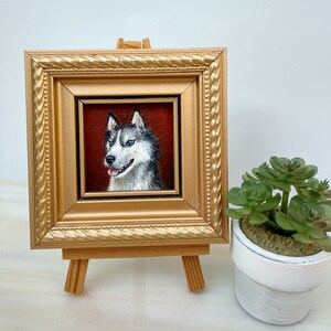 May include: A framed painting of a husky dog with blue eyes. The painting is in a gold frame and is on a small wooden easel.