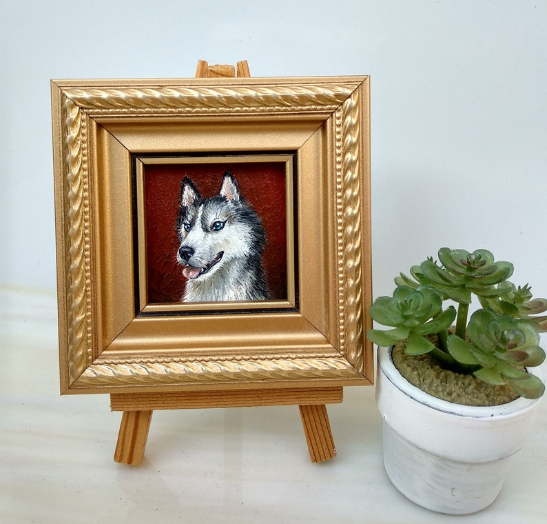 May include: A framed painting of a husky dog with blue eyes. The painting is in a gold frame and is on a small wooden easel.