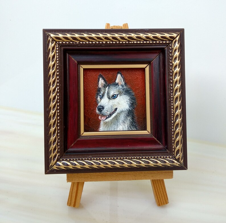 May include: A framed painting of a white and grey husky dog with blue eyes. The dog is looking to the right and has its tongue slightly sticking out. The painting is in a dark wood frame with a gold trim. The frame is on a small wooden easel.