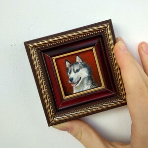 May include: A framed miniature painting of a husky dog with blue eyes. The dog is painted in a realistic style with white fur and black markings. The frame is brown with gold accents.