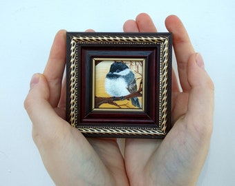 May include: A small framed painting of a black-capped chickadee perched on a branch. The bird is painted in realistic detail with a white background.