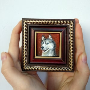 May include: A framed miniature painting of a husky dog with blue eyes. The dog is painted in a realistic style with white fur and black markings. The frame is a dark wood with a gold trim.